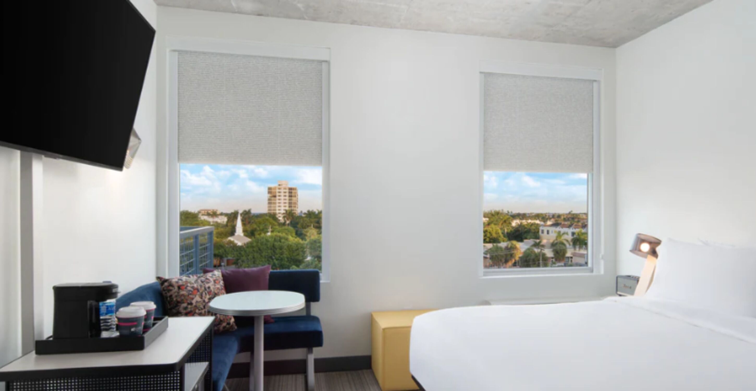 A hotel room with a bed, a small table with a chair, a TV, and two windows with blinds.