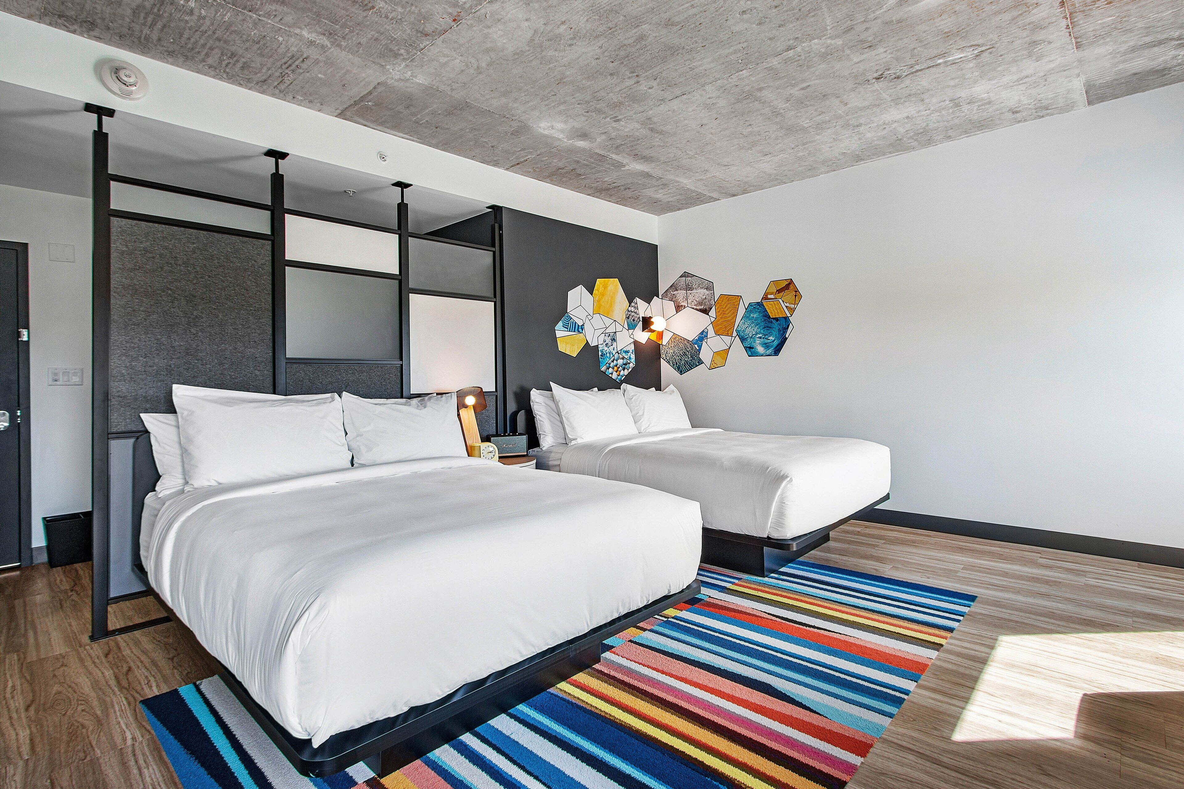 A modern hotel room with two beds, a colorful striped rug, and a unique hexagonal wall art piece.