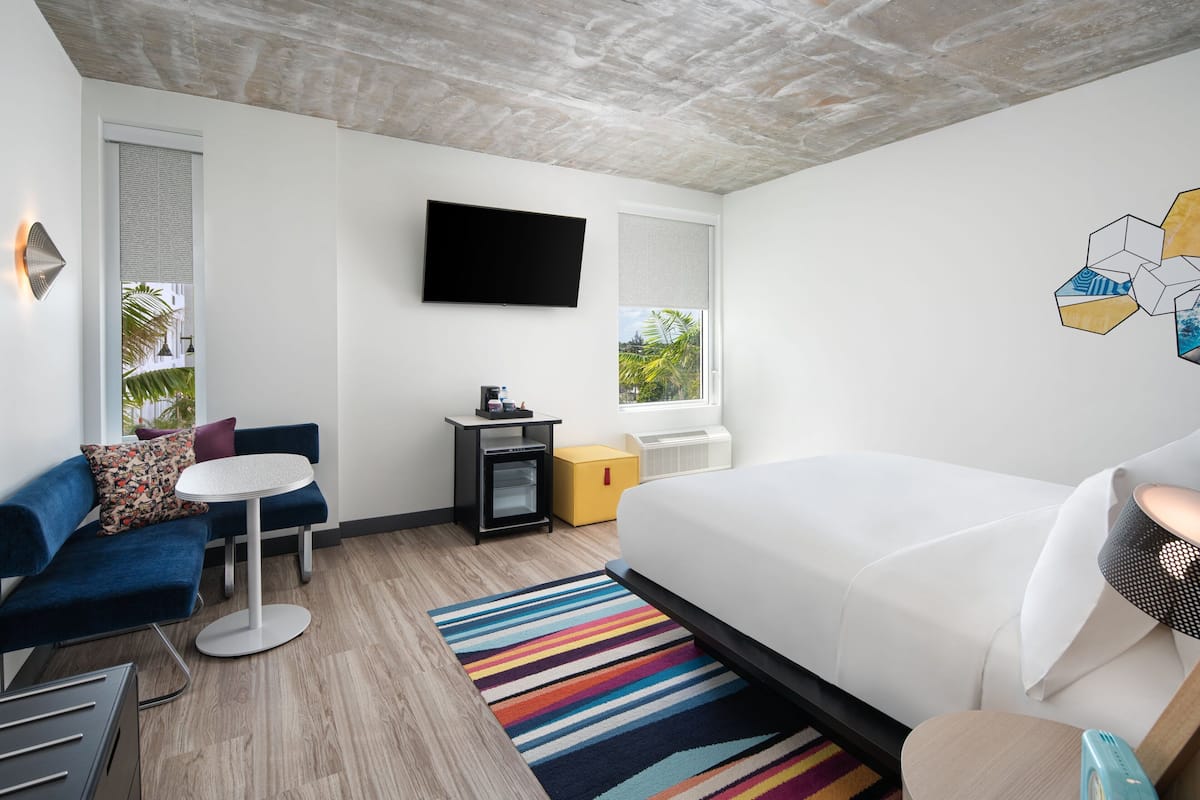 A modern hotel room with a large bed, a blue sofa, a small table, a flat-screen TV, and a window with a view of palm trees.