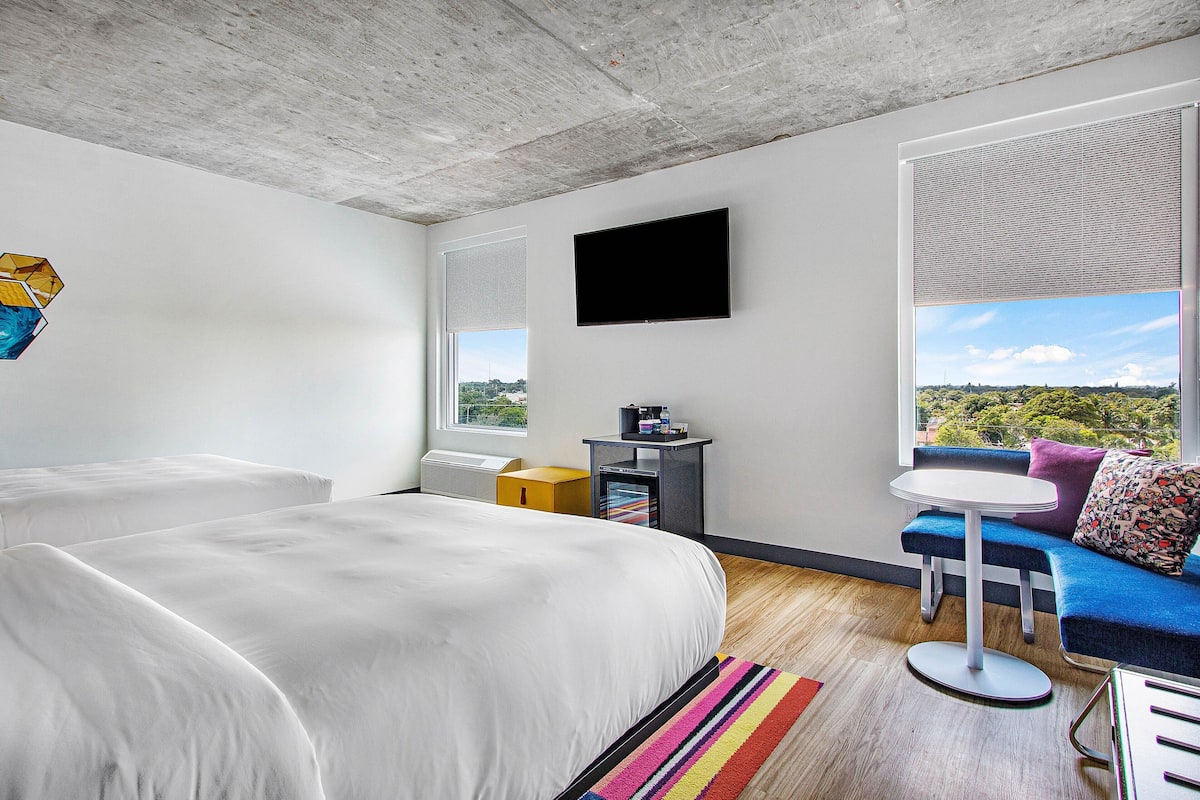 A modern hotel room with a large bed, a small table, a sofa, and a TV on the wall.