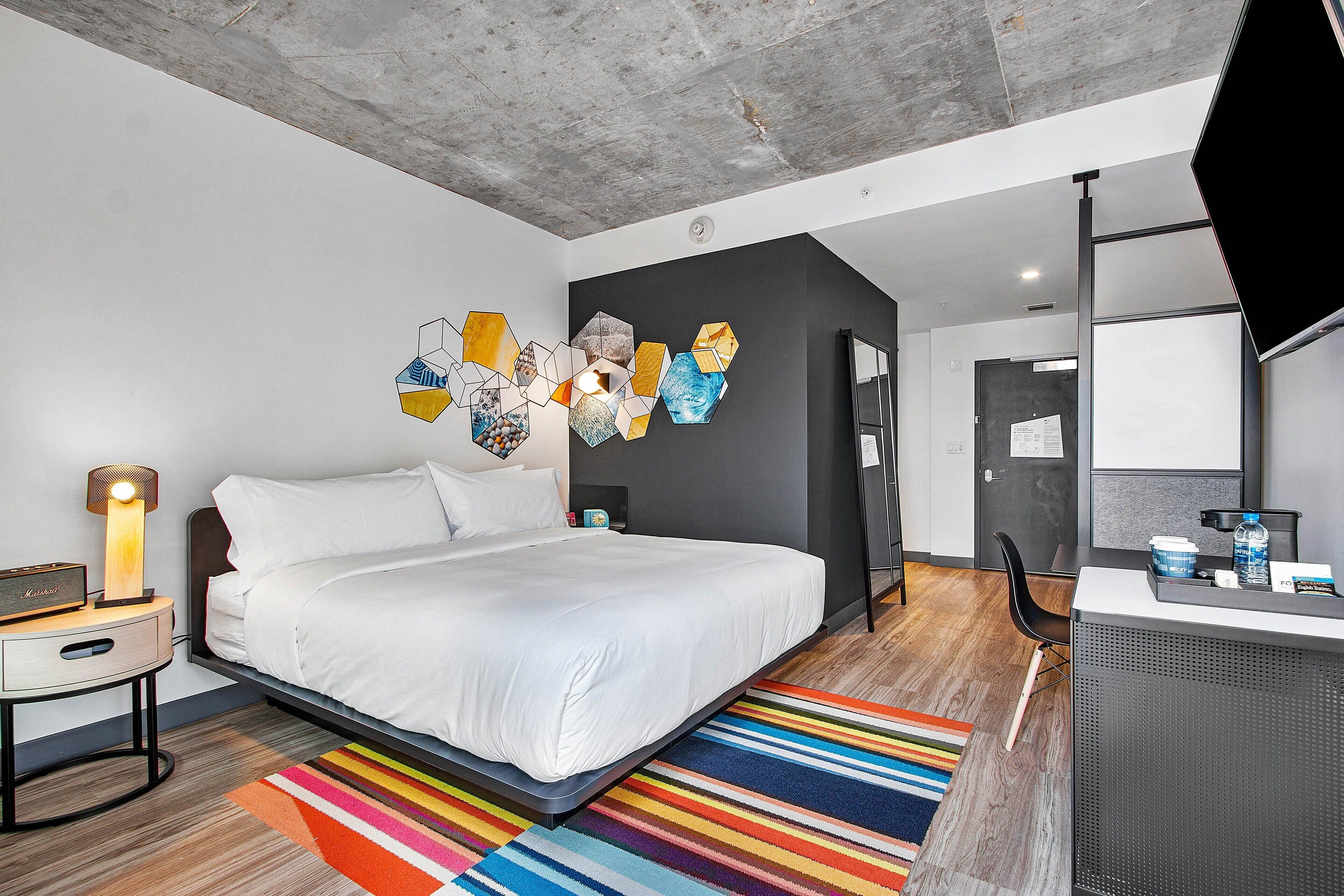 Ett modernt hotellrum med en säng, ett sängbord, en lampa och en färgglad, randig matta.