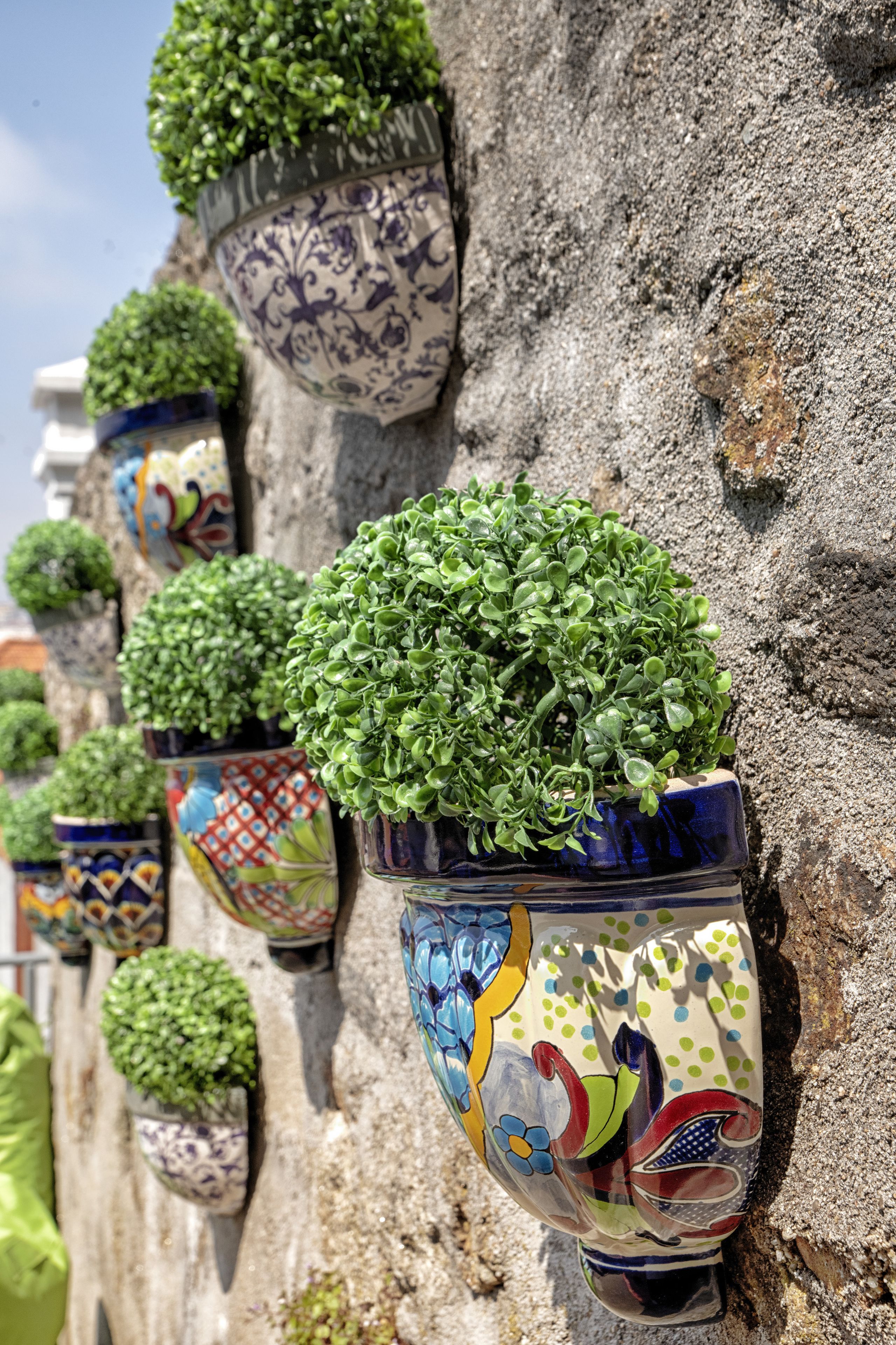 Una hilera de macetas de cerámica coloridas con plantas verdes, montadas sobre una pared de piedra texturizada.