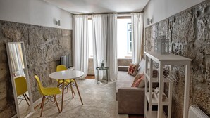 Un salón acogedor con pared de piedra, una mesa redonda con sillas amarillas, un sofá con cojines estampados y un mueble blanco para estanterías.