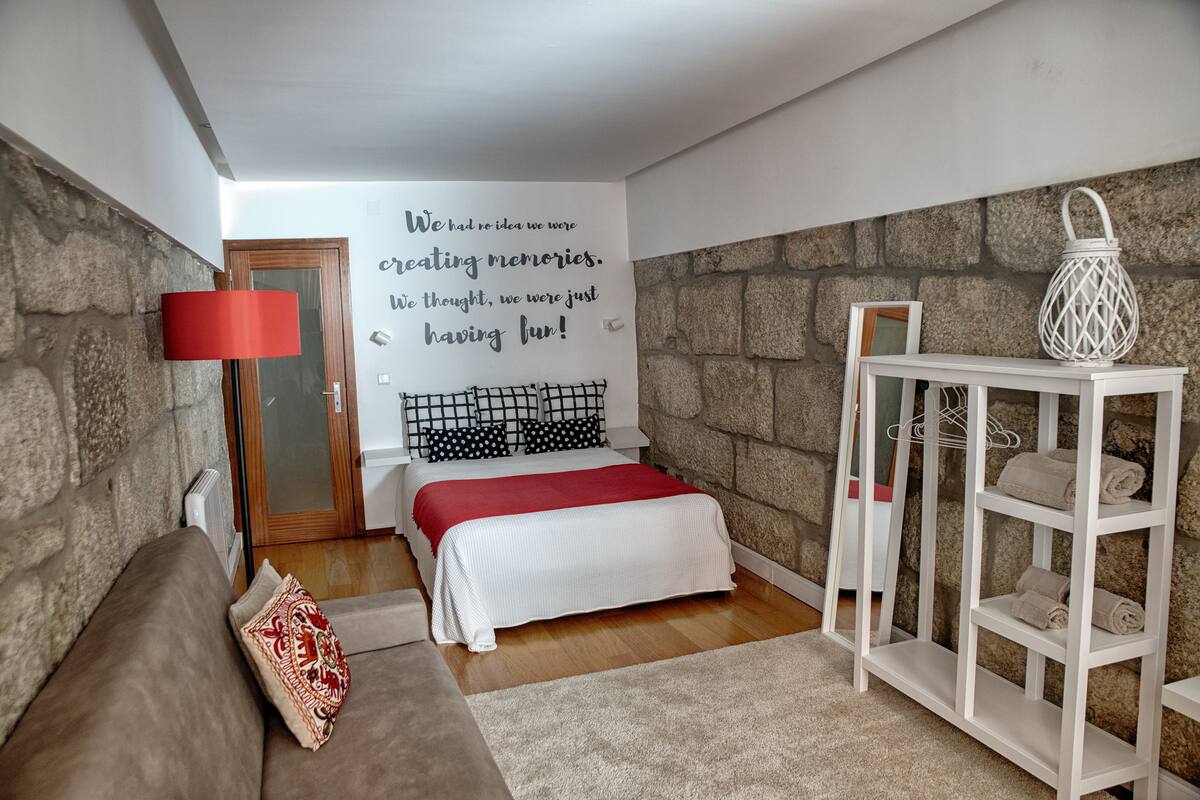 A cozy bedroom with a stone wall, a bed with a red blanket, a white bedside table, and a red floor lamp.