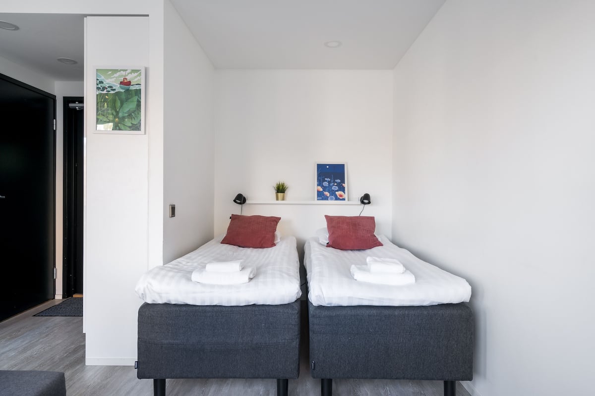 A modern room with two single beds, a small shelf with decorative items, and a door leading to another room.