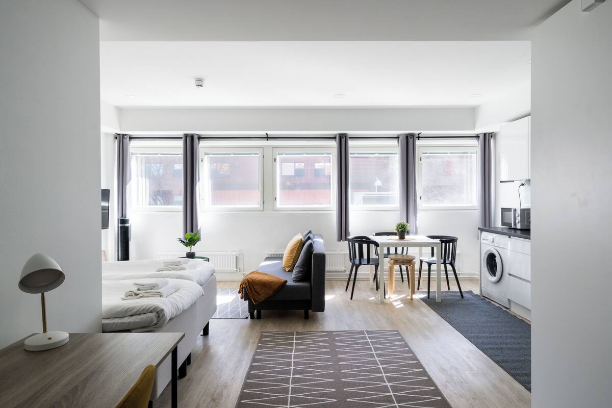 A modern, well-lit room with a sofa, a dining table, a washing machine, and a large window with curtains.