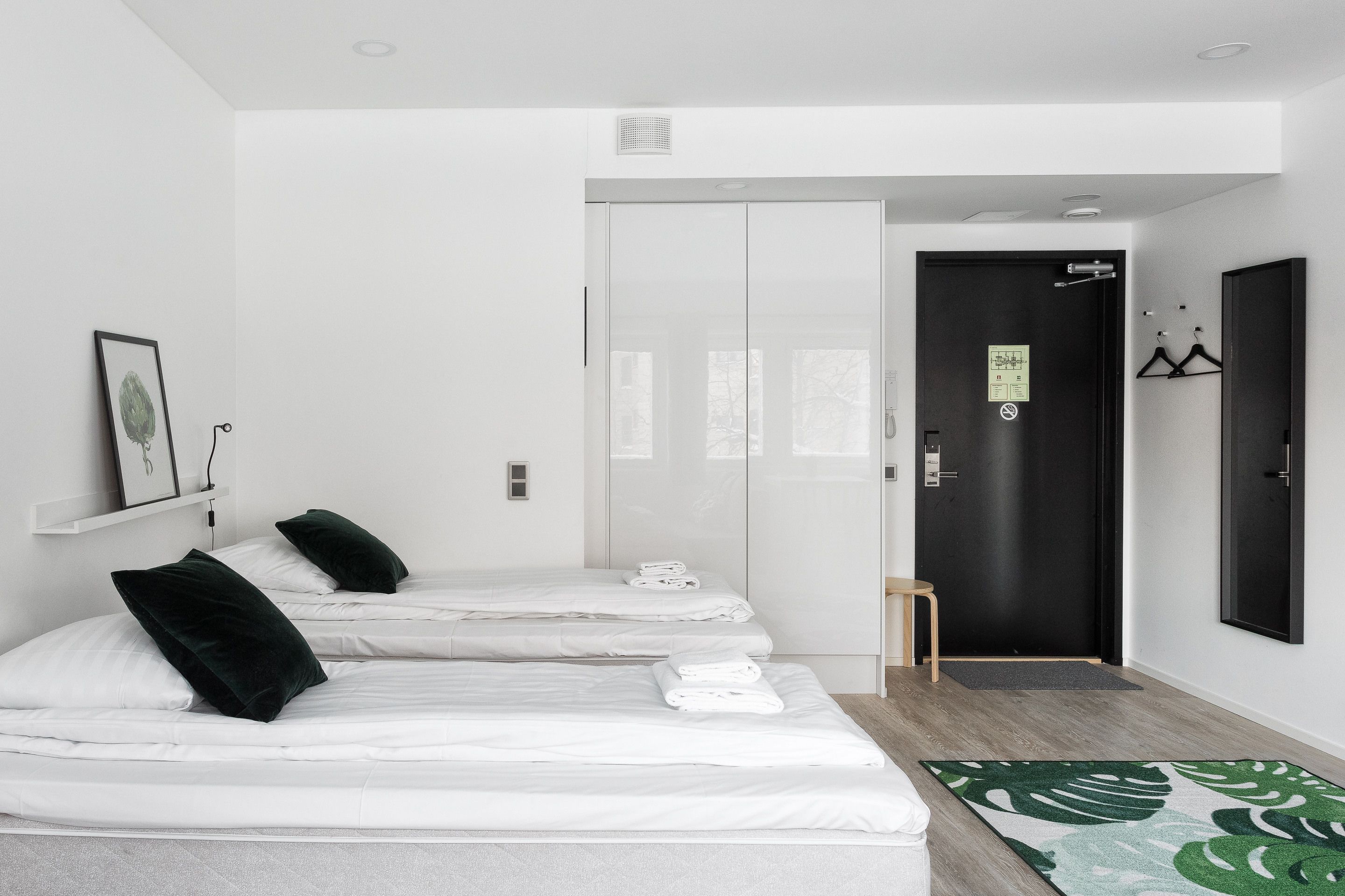 Une chambre d’hôtel avec trois lits, une porte noire, un petit tabouret en bois et un tapis à motifs verts.