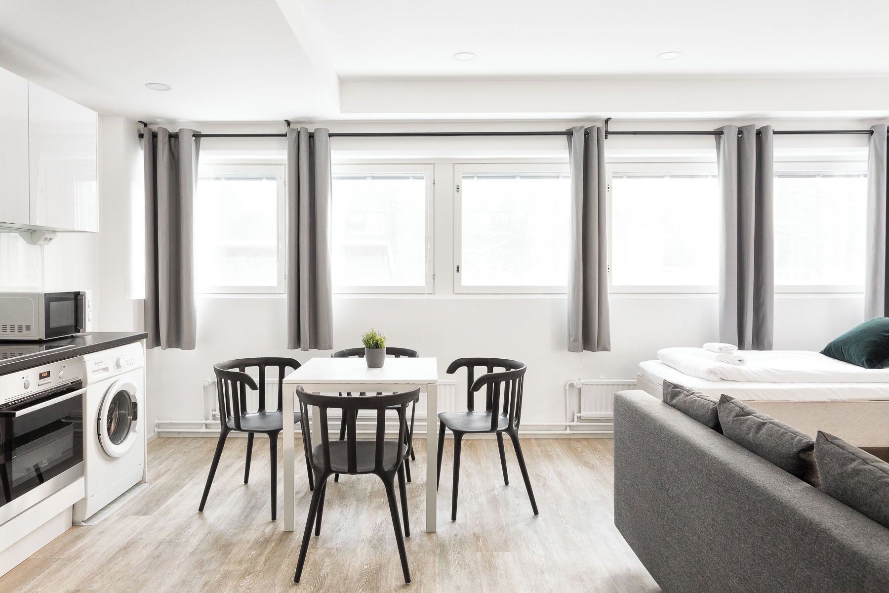 Een moderne keuken met een magnetron, oven en wasmachine; een eethoek met een tafel en stoelen; een grijze bank; en grote ramen met grijze gordijnen.