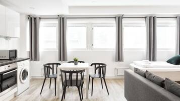 A modern kitchen with a microwave, oven, and washing machine; a dining area with a table and chairs; a grey sofa; and large windows with grey curtains.