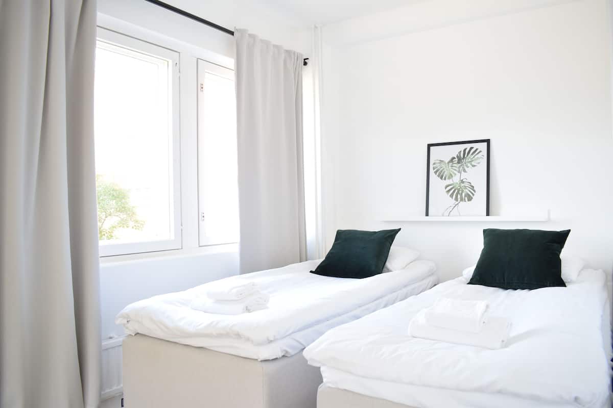A hotel room with two single beds, white bedding, a window with curtains, and a framed plant on the wall.
