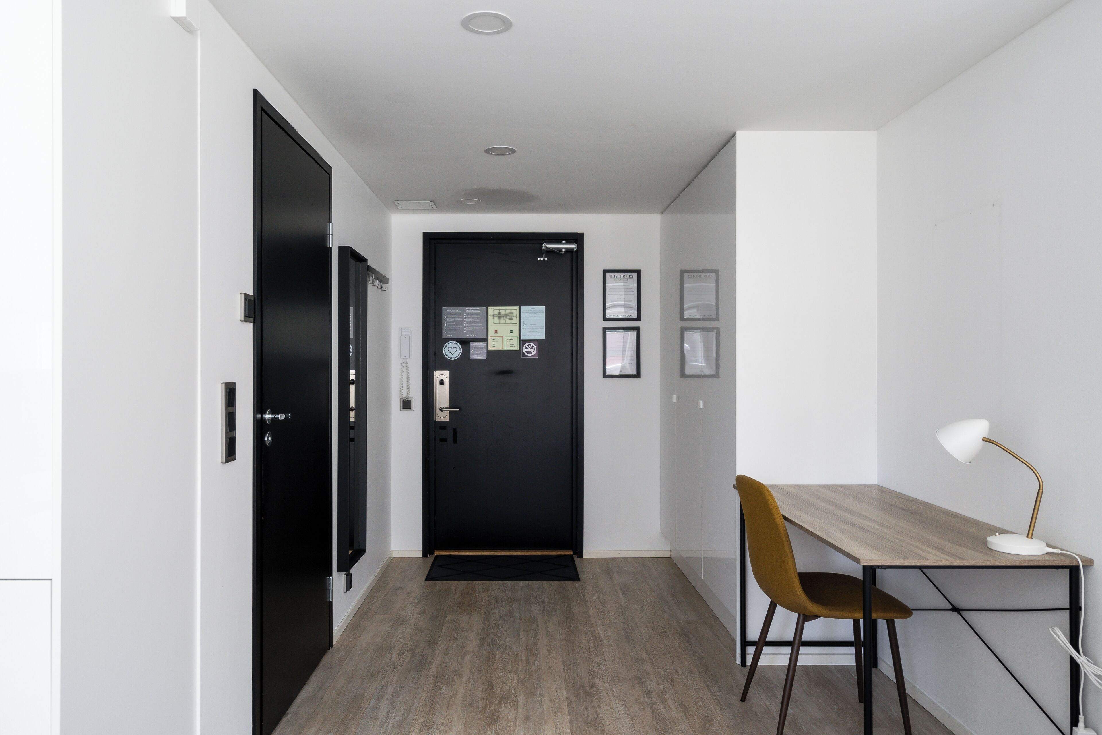A hallway with a black door, a desk with a chair, and a white desk lamp.