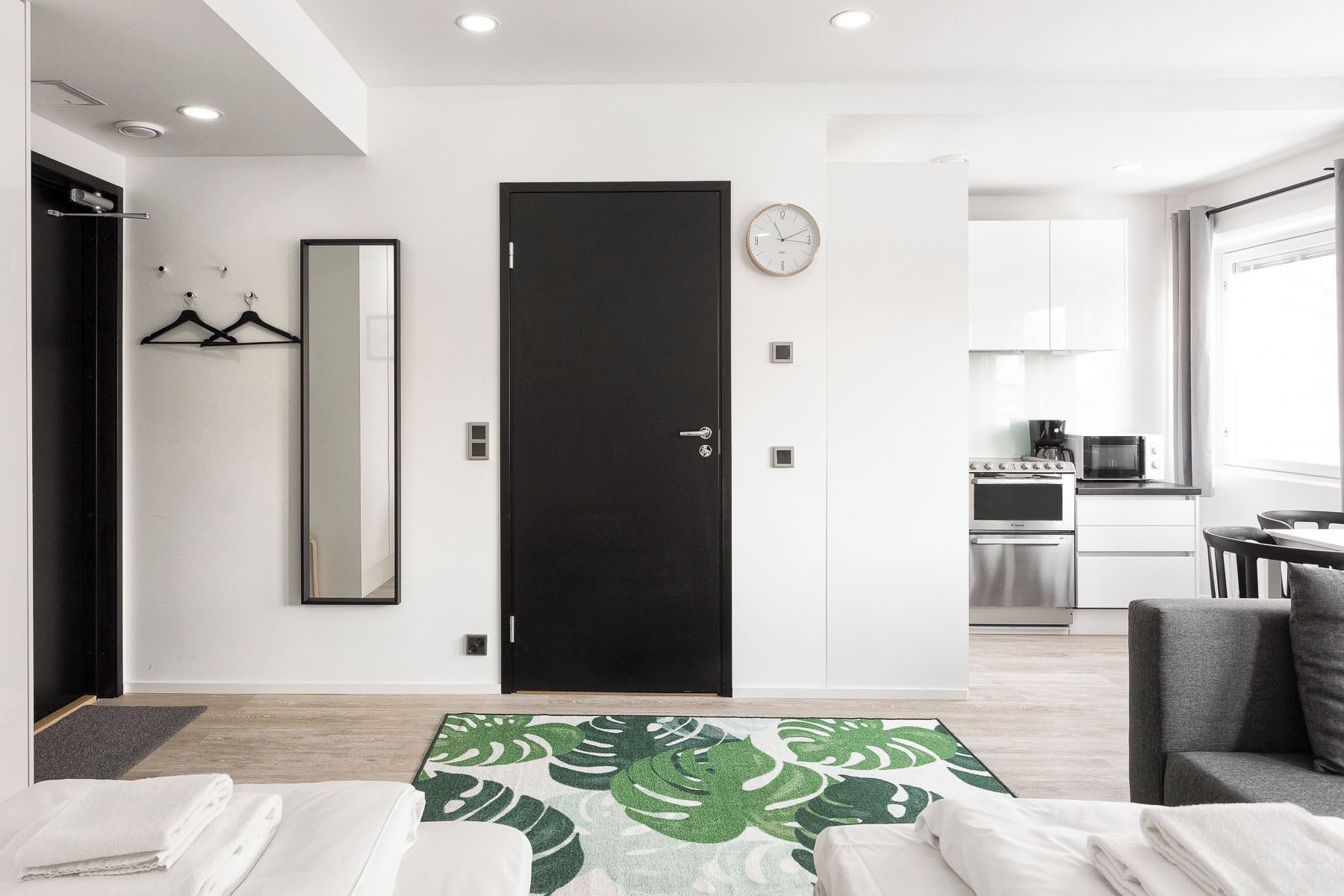 Habitación de hotel moderna con una cama, una puerta negra, un espejo, un reloj, un kitchenette y vistas al exterior.