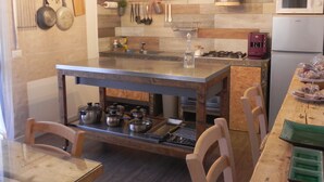 A modern kitchen with a central island, stainless steel appliances, hanging utensils, and a wooden dining table with chairs. - Gea apartment Etna (Nicolosi)