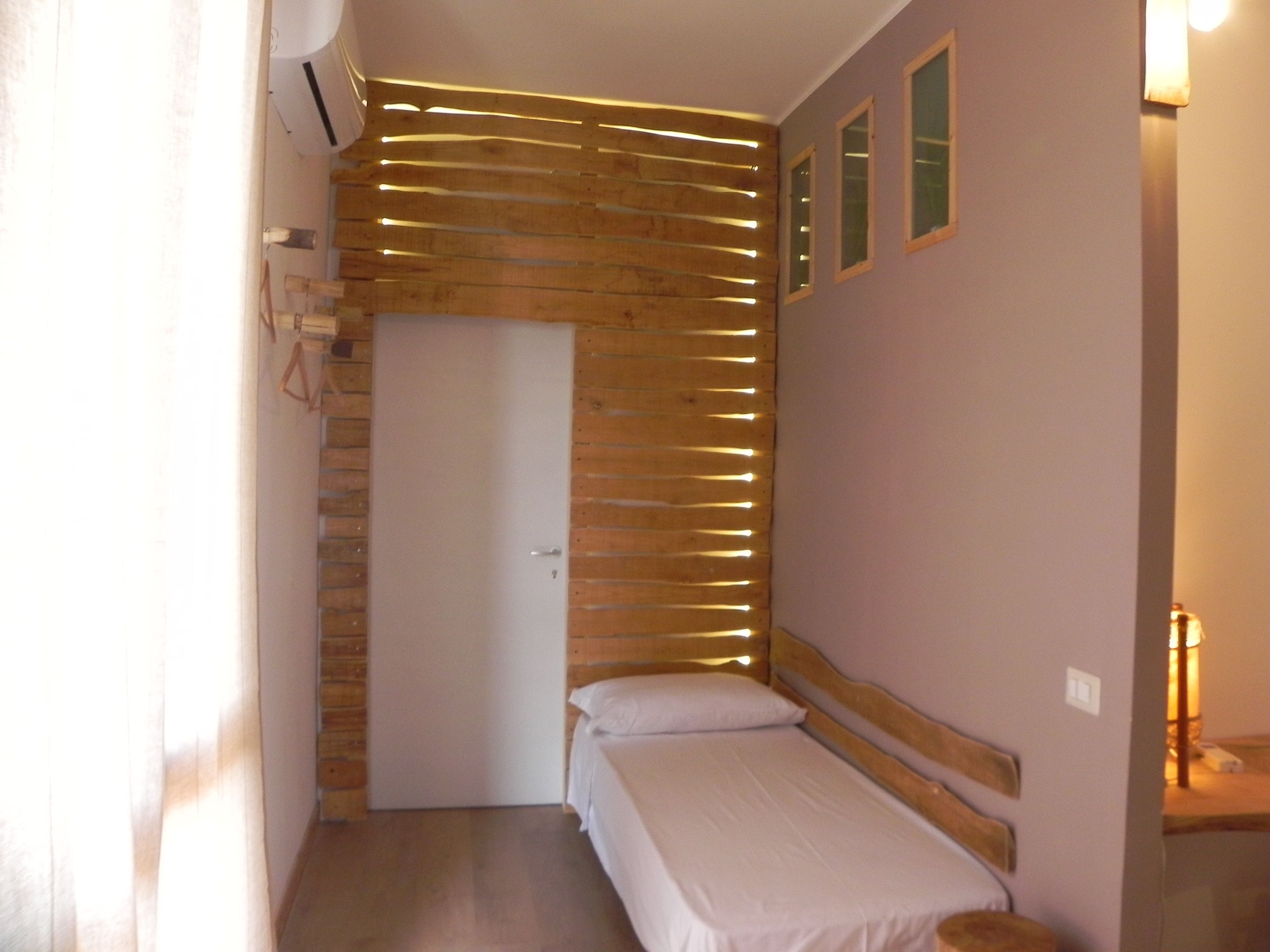 A small, compact room with a wooden slatted wall, a single bed, a door, and a small table with a lamp.