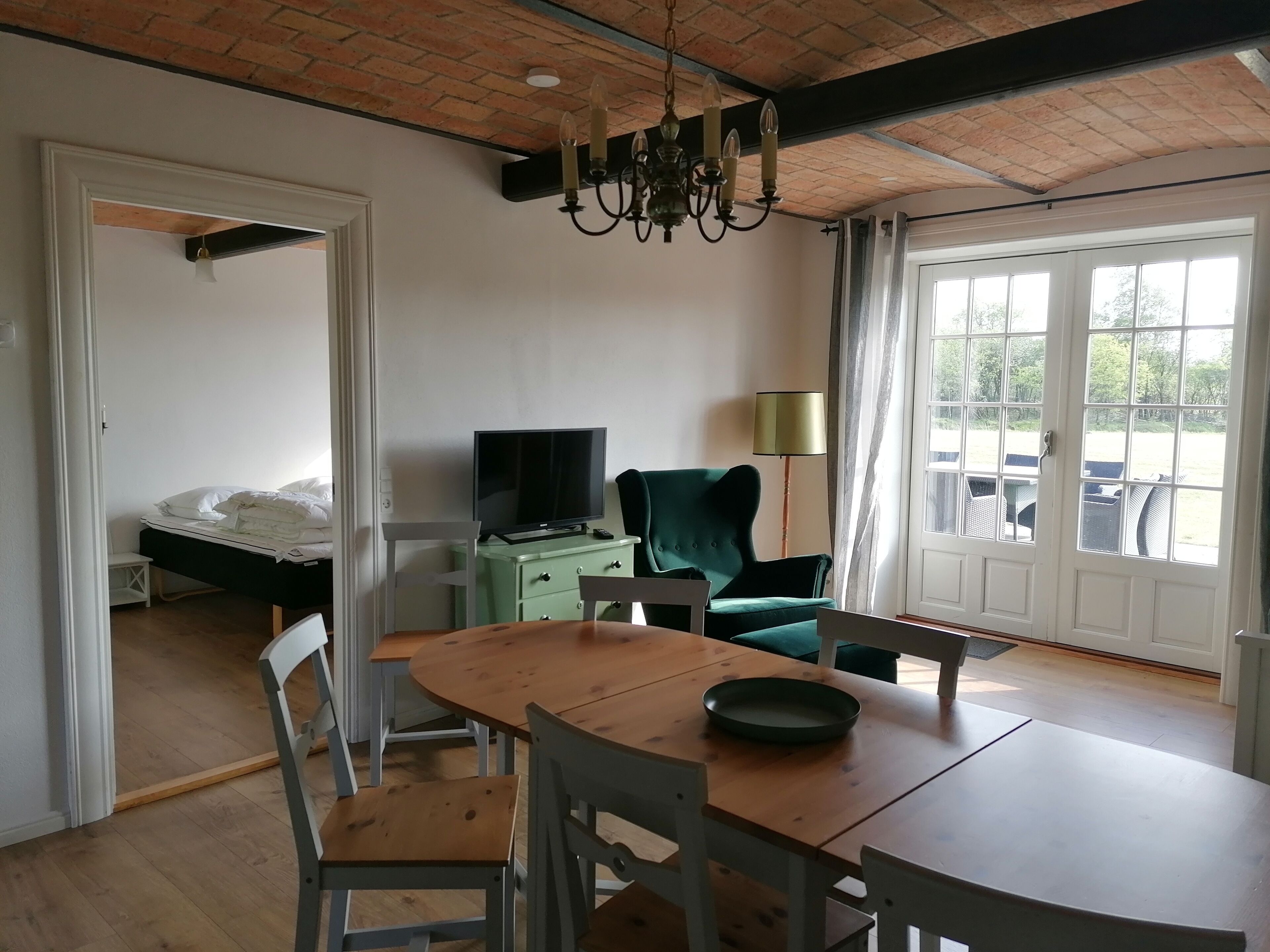 Una habitación con una mesa de comedor de madera, sillas, un televisor sobre un mueble verde, un sillón verde y un ventanal con vistas a muebles de exterior.