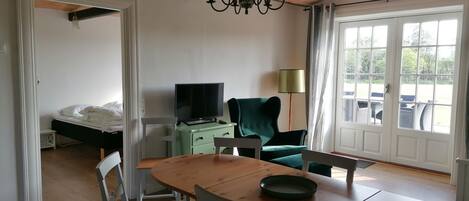 A room with a wooden dining table, chairs, a television on a green cabinet, a green armchair, and a large window with a view of outdoor furniture.