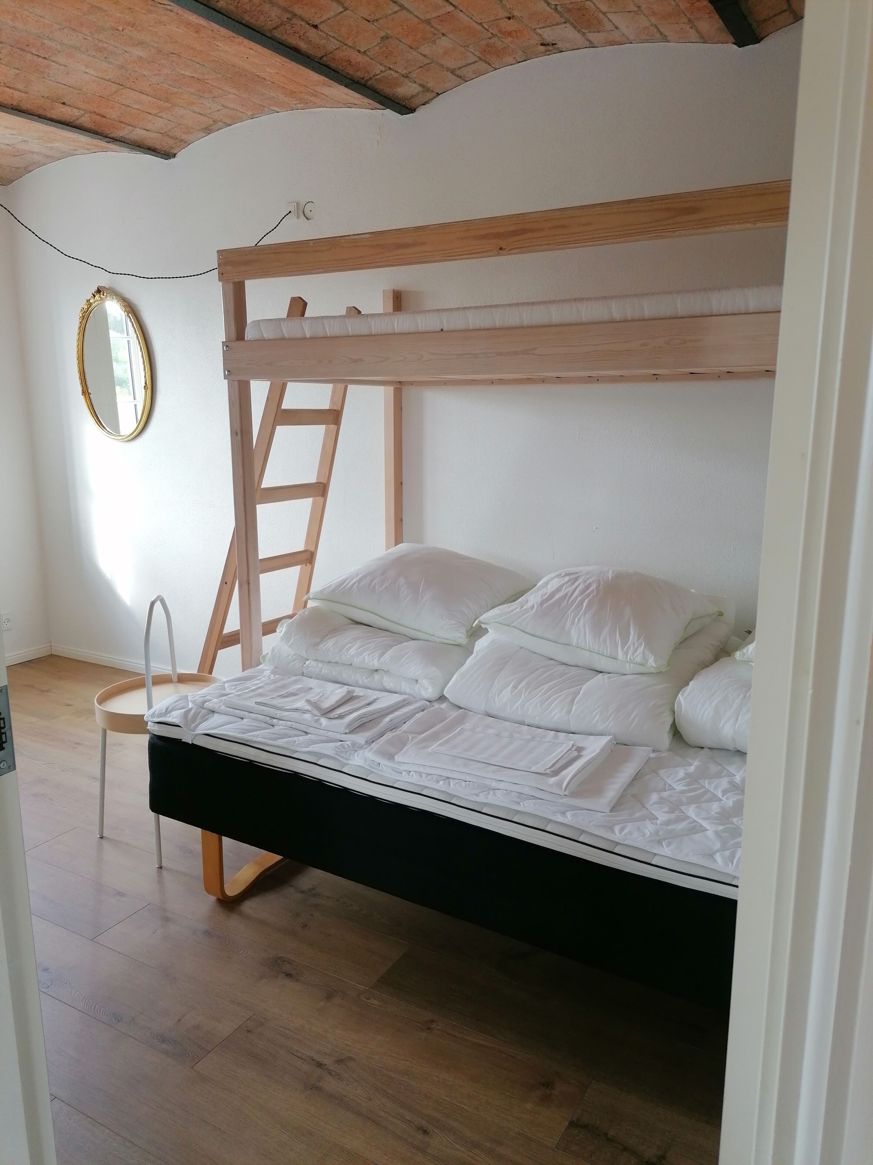 Una cama litera con ropa de cama blanca, una escalera de madera, un espejo redondo pequeño y una silla de madera.