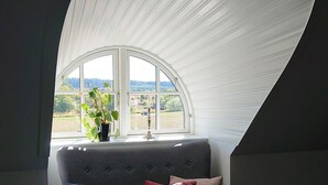 Una habitación con techo curvo, ventana con vista a un paisaje verde, sofá gris con un cojín rojo y una colcha acolchada oscura.