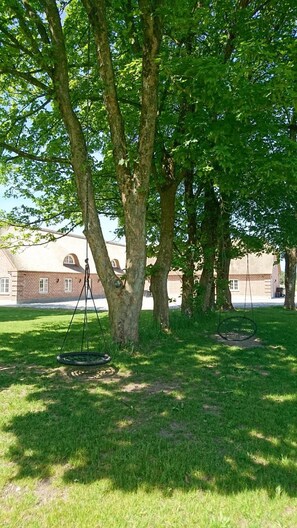 A grassy area with two swings under large trees, a brick building in the background. - Aagaarden (Billund)