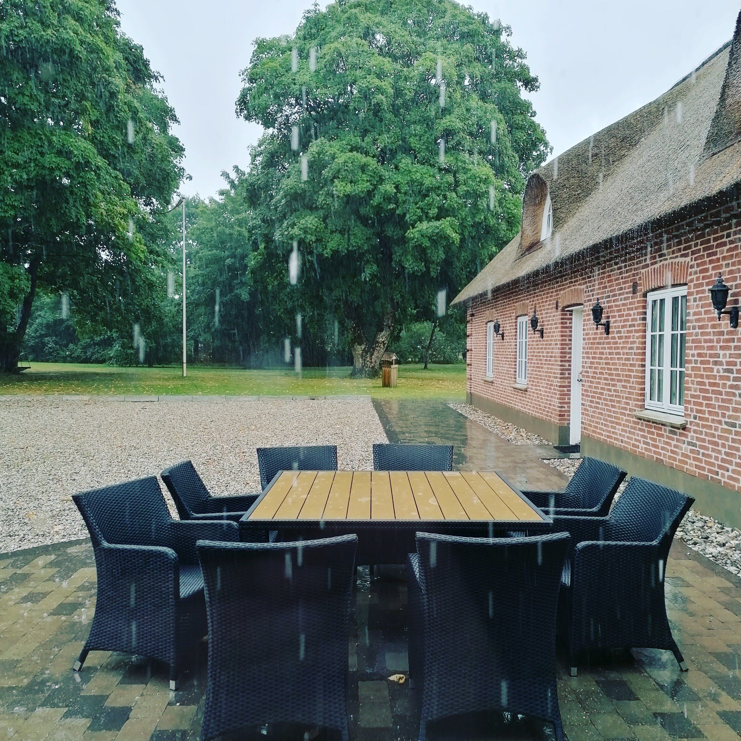 A patio with a wooden table and wicker chairs, a brick building with a thatched roof, and a gravel area.
