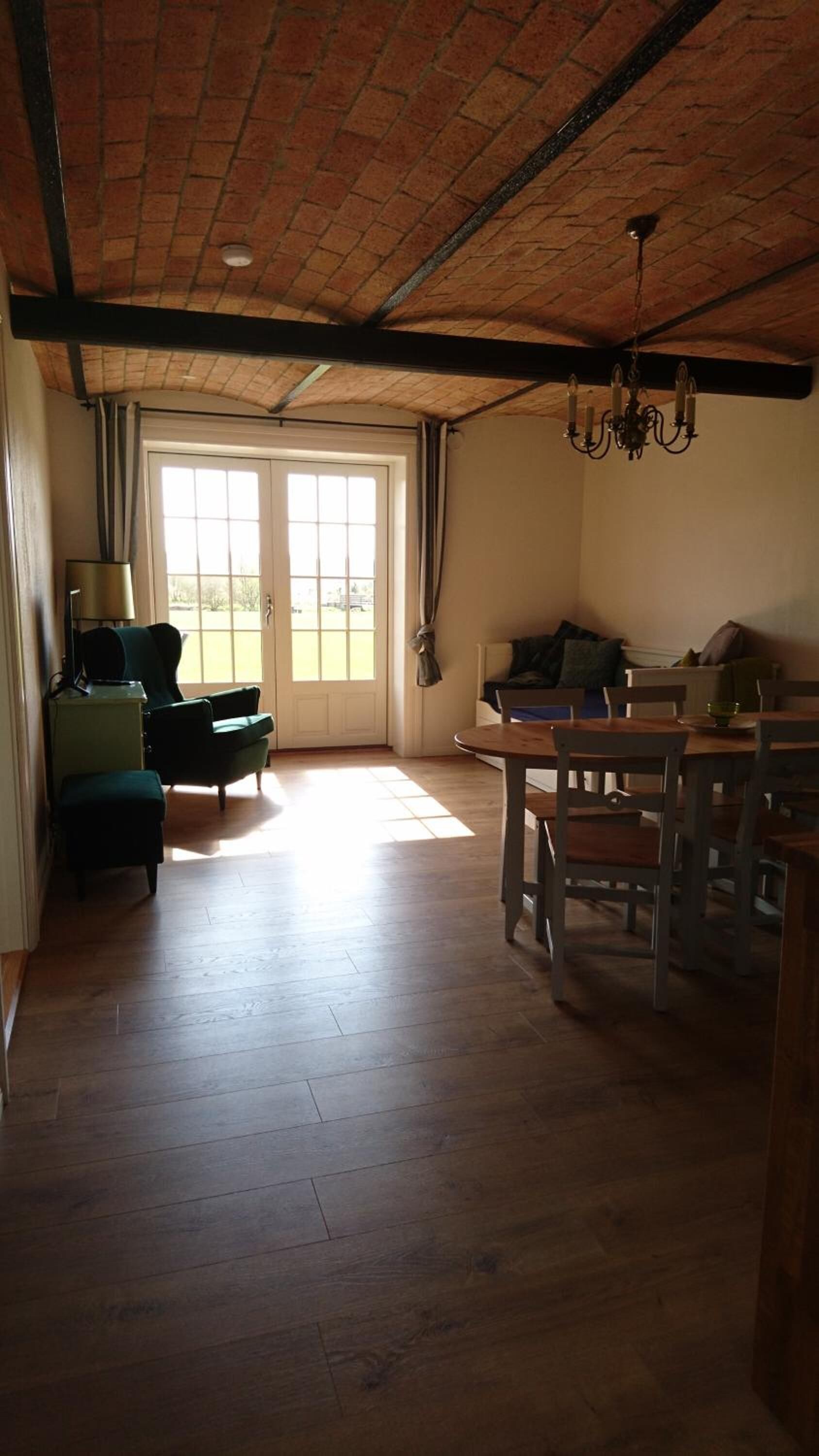 A spacious living room with a high ceiling, a dining area with wooden furniture, and a large sliding glass door leading to a bright outdoor area.