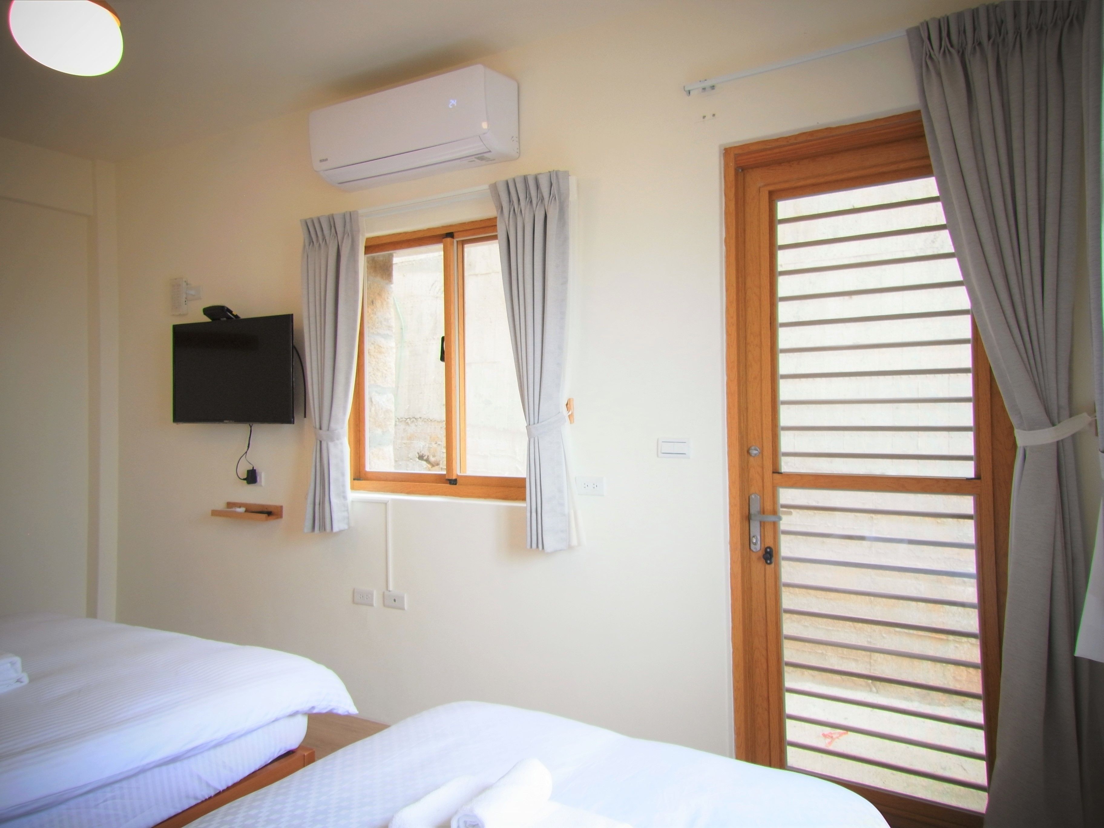 A room with two beds, an air conditioning unit, a television, a window with curtains, and a door with blinds.