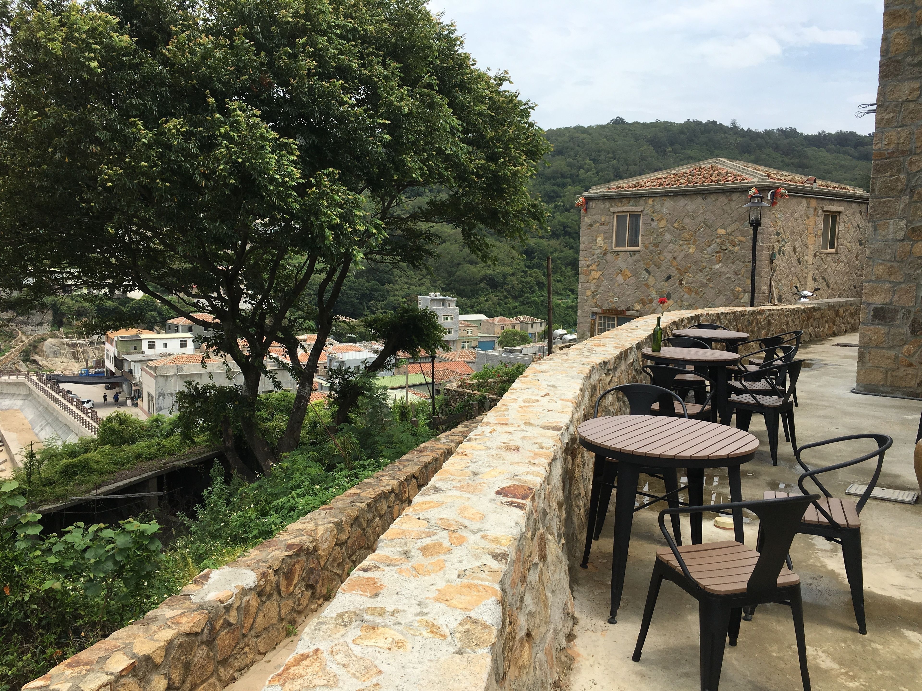 A stone-walled outdoor seating area with tables and chairs, overlooking a hillside with buildings and trees.