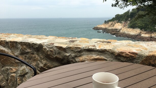 A white mug on a brown table with a view of the ocean and rocky cliffs. - Mulight B&B (Nangan)