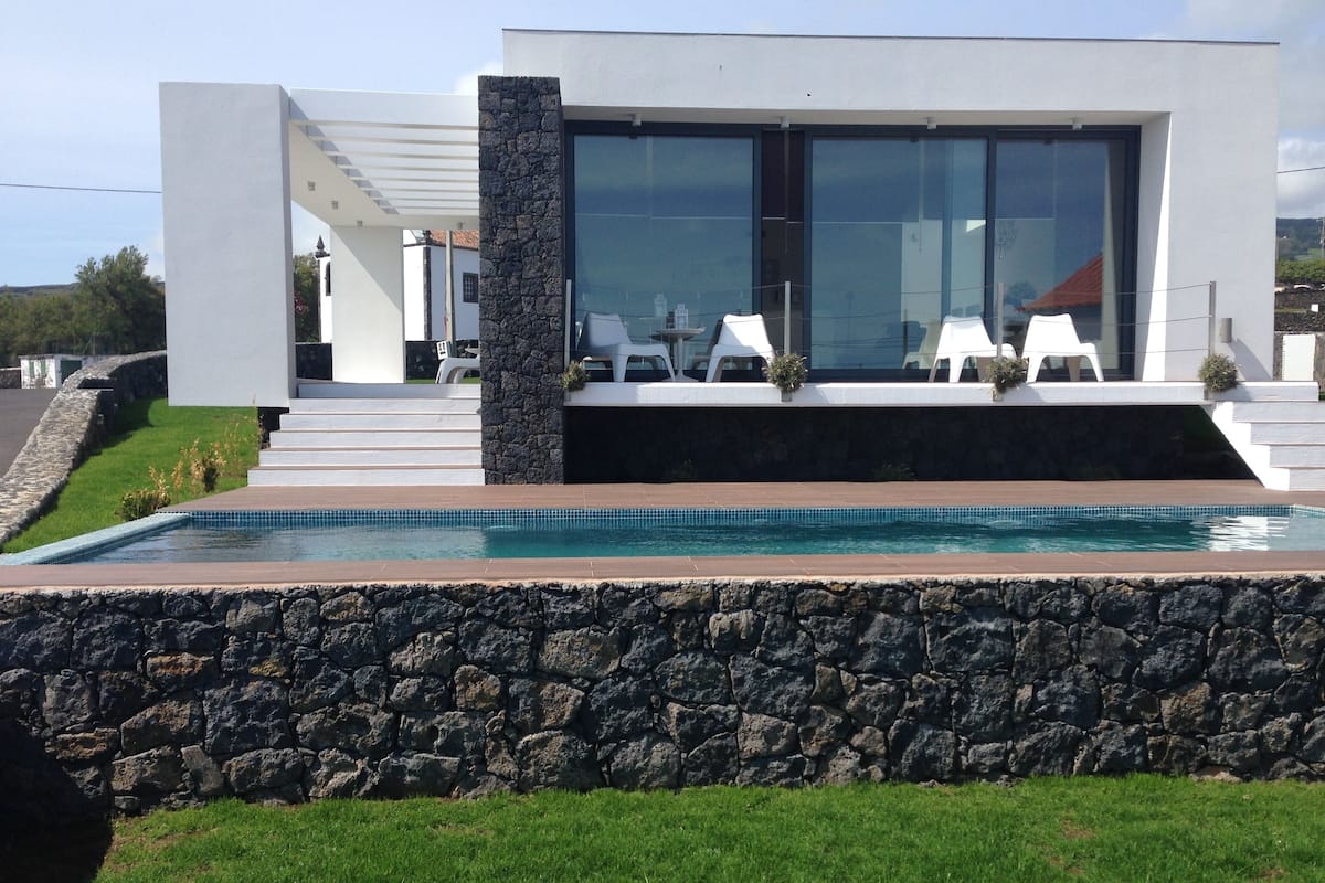 A modern house with a swimming pool, stone walls, and a patio with white chairs.