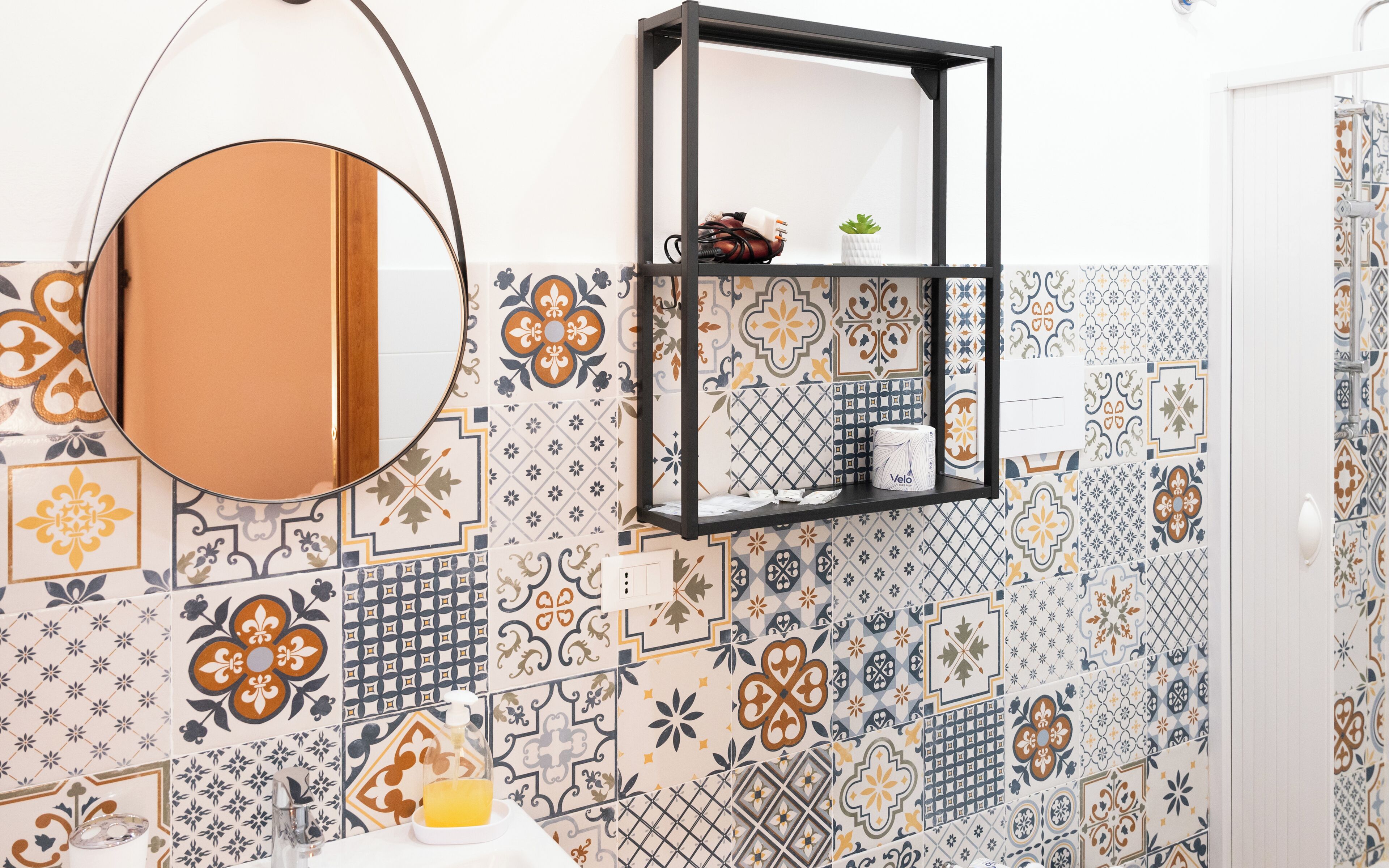 A bathroom with a round mirror, a shelf with toiletries, and a patterned tile wall.