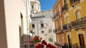 Un vicolo stretto con edifici su entrambi i lati, un'auto bianca parcheggiata a destra e un balcone fiorito di fiori rossi.