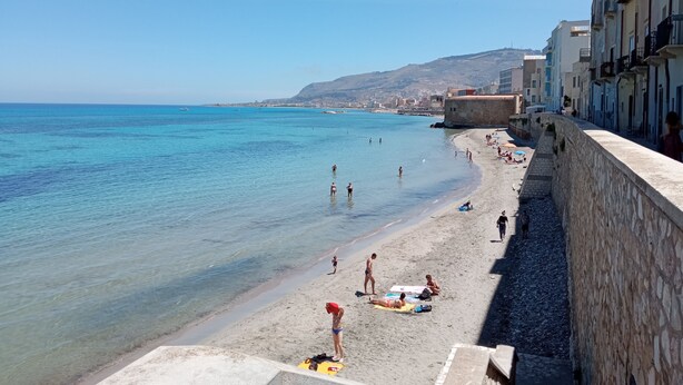 Una spiaggia costiera con acque blu cristalline, persone che prendono il sole e edifici lungo la riva.