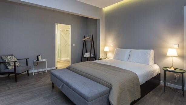 Una habitación de hotel moderna con una cama grande, lámparas de noche, un banco, una mesita y un vestidor con ducha.