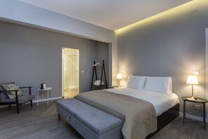Una habitación de hotel moderna con una cama grande, lámparas de noche, un banco, una mesita y un vestidor con ducha.
