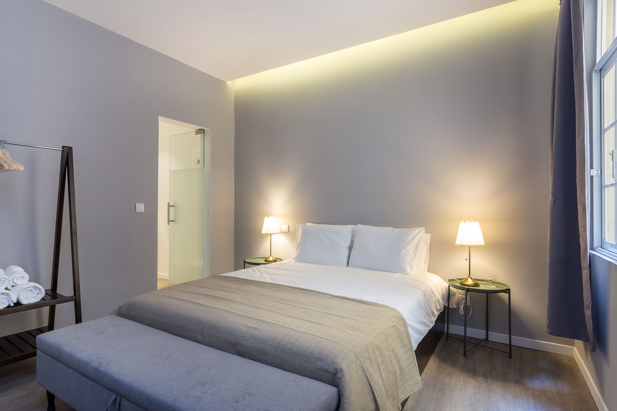 A modern hotel room with a large bed, two bedside tables with lamps, a bench, a towel rack, and a glass shower door.