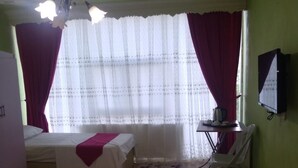 A hotel room with two beds, a small table with a chair, a television on the wall, and a window with sheer curtains. - Otel Alimoglu (Gelendost)