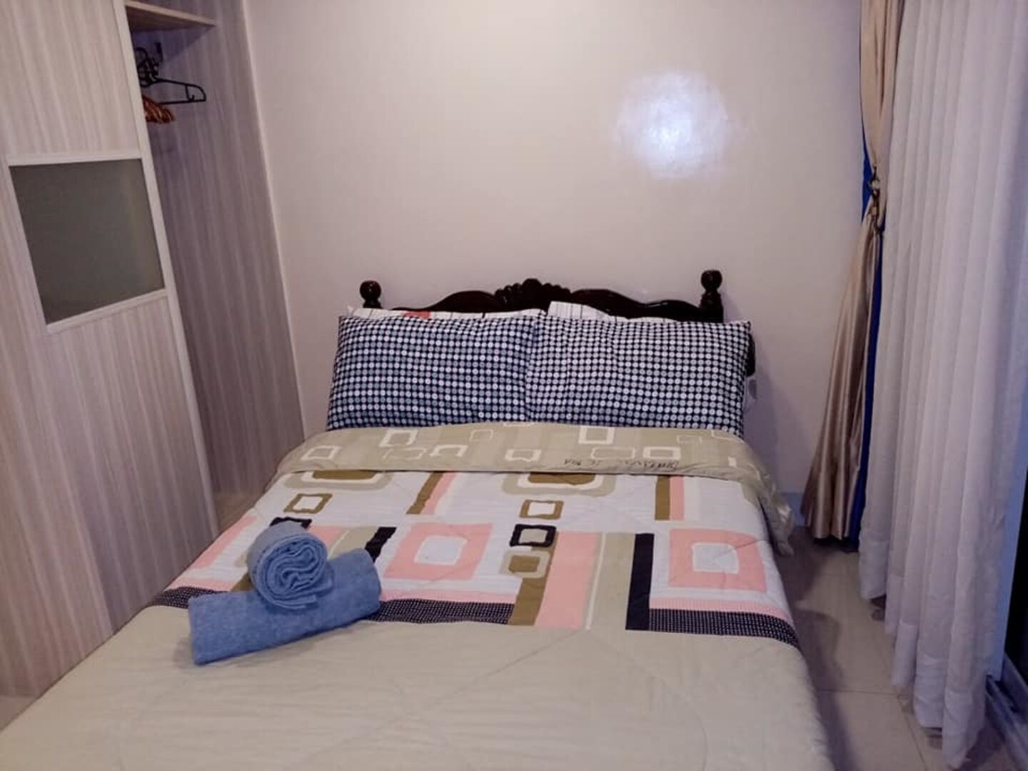 A neatly made bed with a patterned bedspread and two pillows, a blue towel, and a small shelf with a mirror and a hanging item.