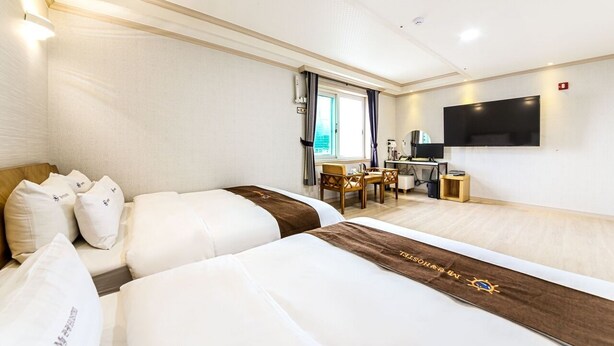 A hotel room with two beds, a desk, a chair, a TV, and a window with curtains. - MJ Tour Hostel (Yeosu)