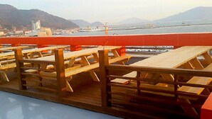 A wooden deck with picnic tables and a red railing overlooking a body of water with distant mountains. - MJ Tour Hostel (Yeosu)