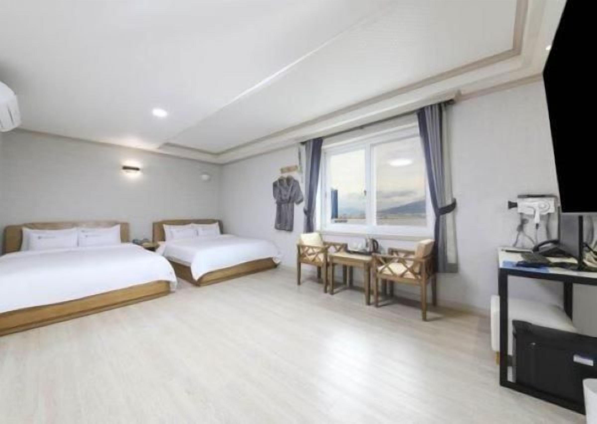 A hotel room with two beds, a desk with a chair, a window with curtains, and a flat-screen TV.