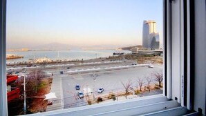 A view from a window overlooking a parking lot with a body of water and a tall building in the distance. - MJ Tour Hostel (Yeosu)