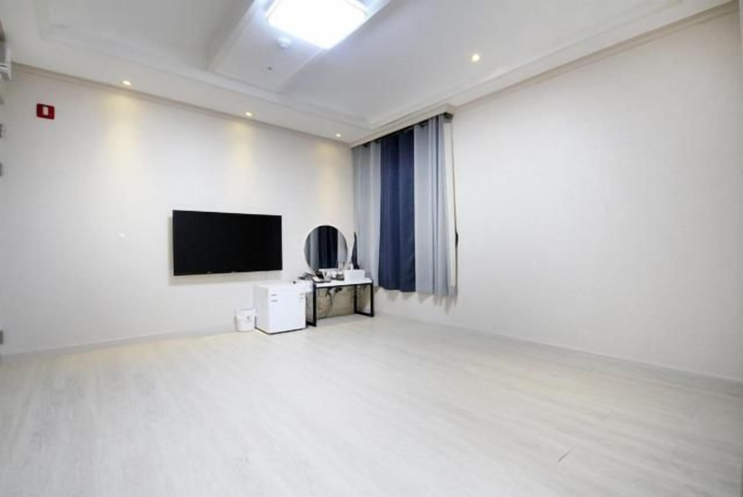 Habitación con televisión de pantalla plana en la pared, un escritorio pequeño con computadora y una ventana con cortinas.