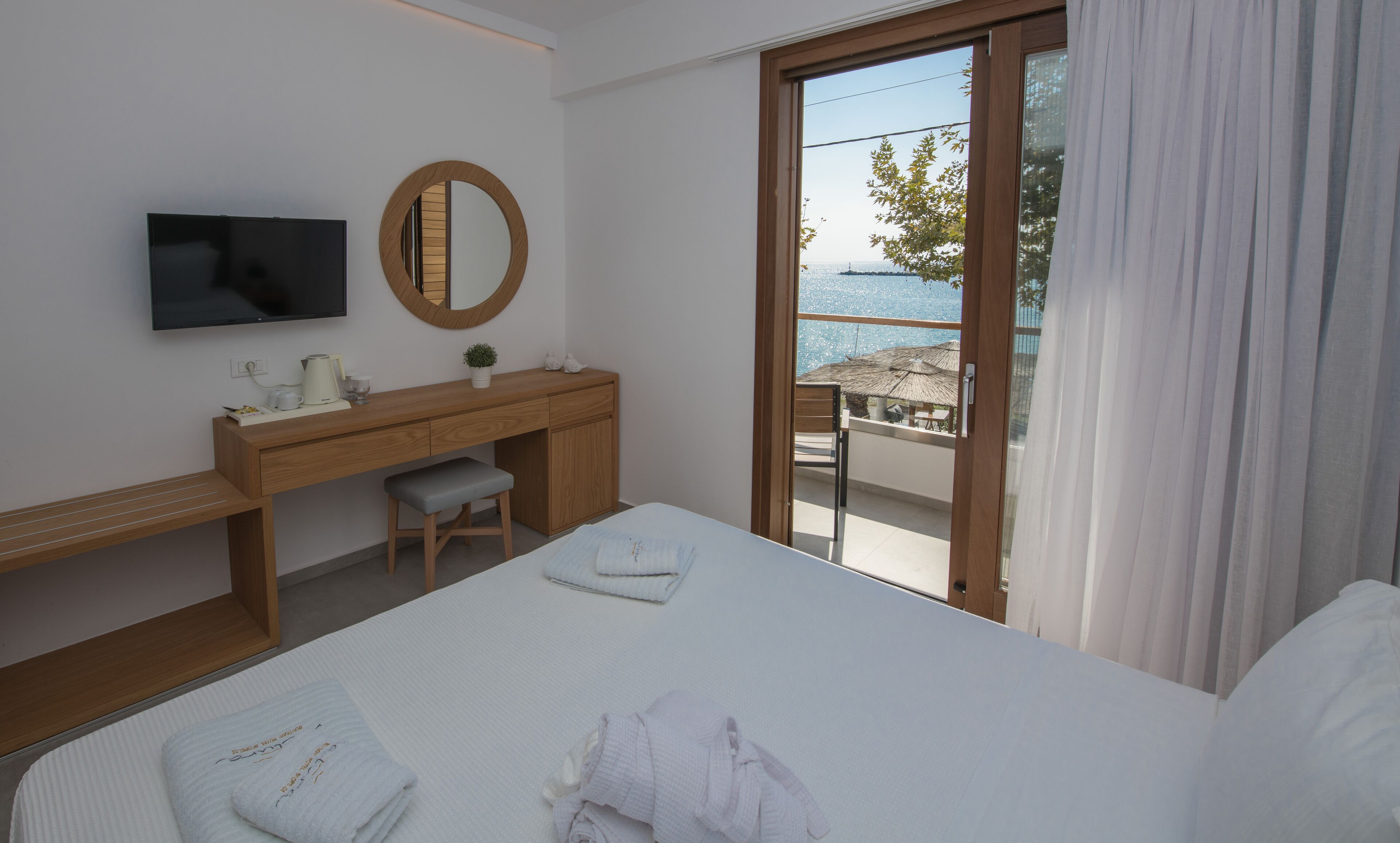 A hotel room with a bed, a desk with a TV, a mirror, and a view of the sea through a sliding door.