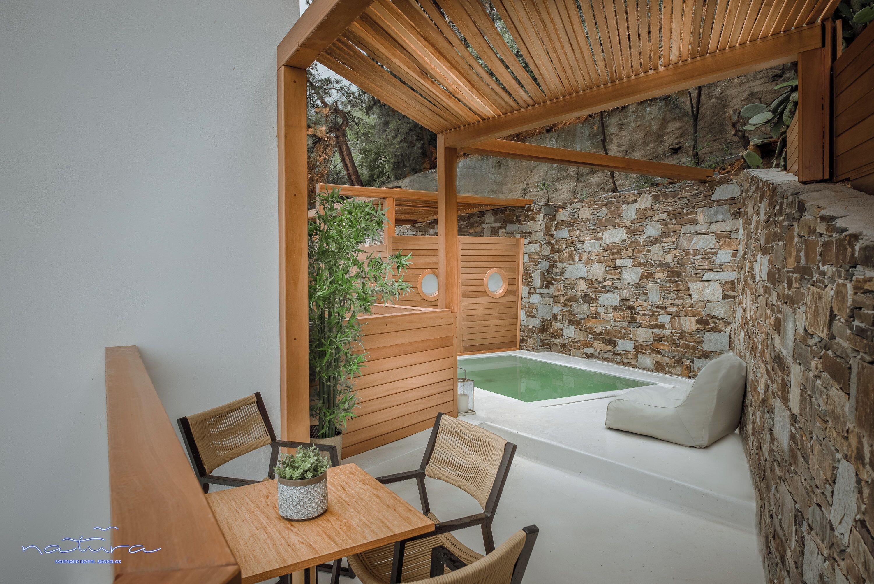 Un área de estar exterior de madera con una mesa y sillas, una pequeña planta en maceta y un muro de piedra de fondo.