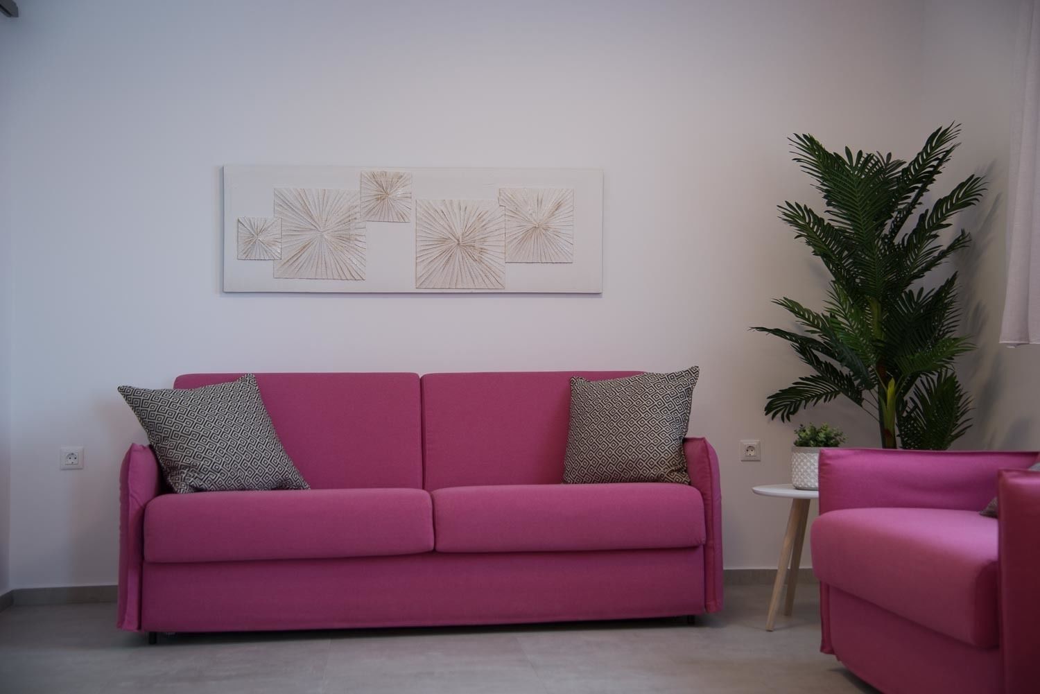 A pink sofa with patterned cushions, a wall-mounted abstract art piece, and a tall green plant.