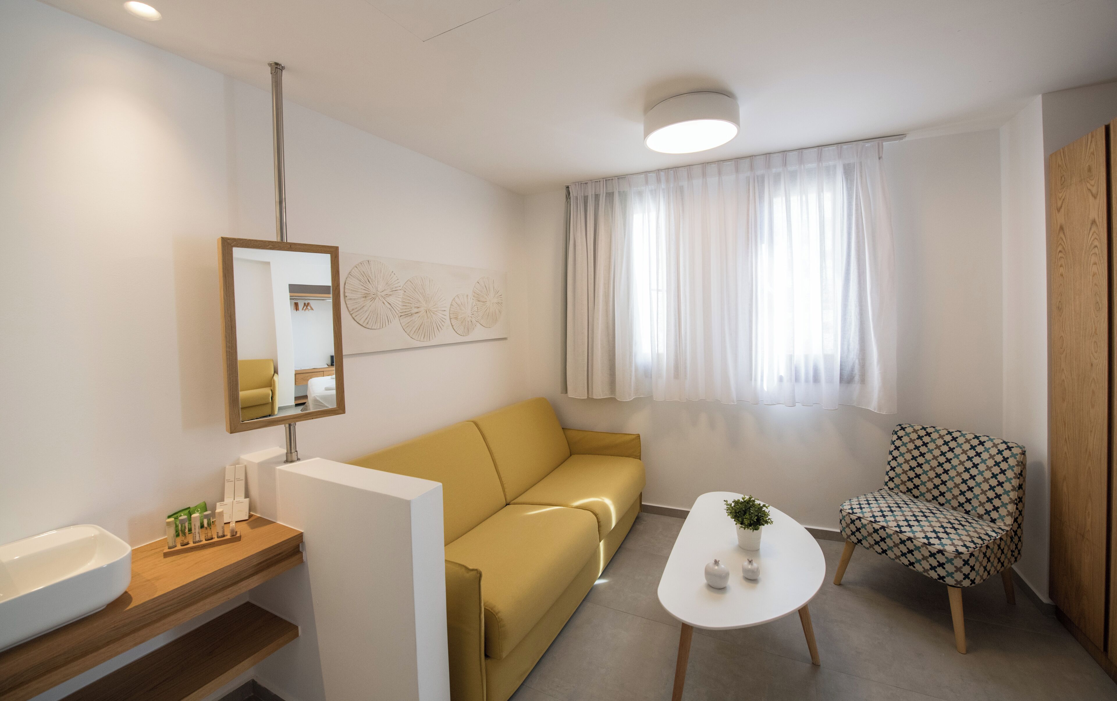 A modern hotel room with a sofa, a small table with a plant, a chair, a sink, and a mirror.