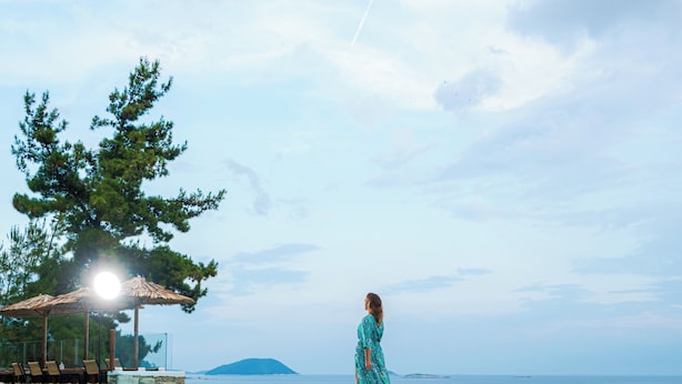 A woman in a blue dress walking along the edge of a pool with a view of the sea and a distant island.