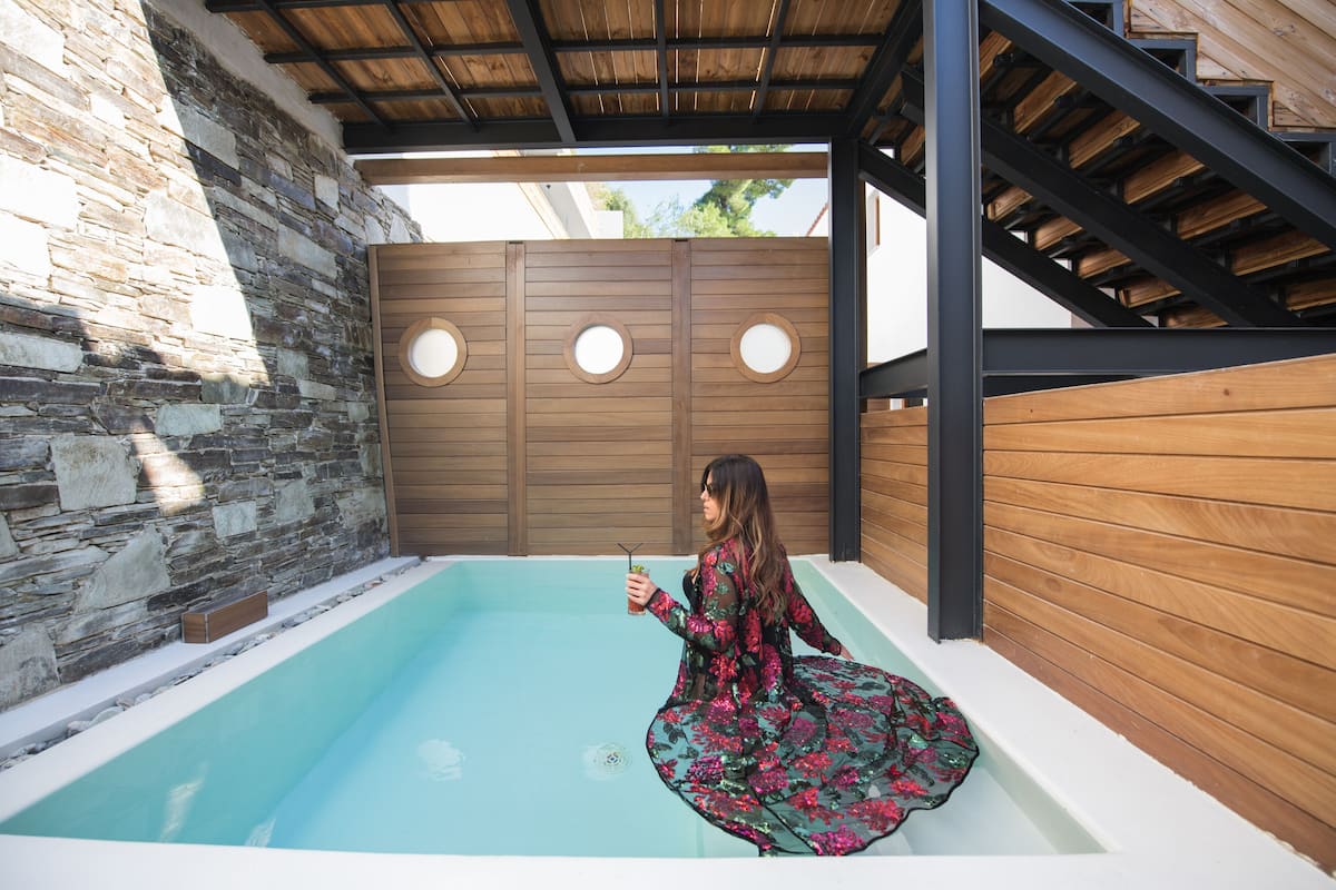 Une femme, vêtue d’une robe à motifs floraux, est assise au bord d’une piscine dont les murs sont en pierre et dont le revêtement est en bois.