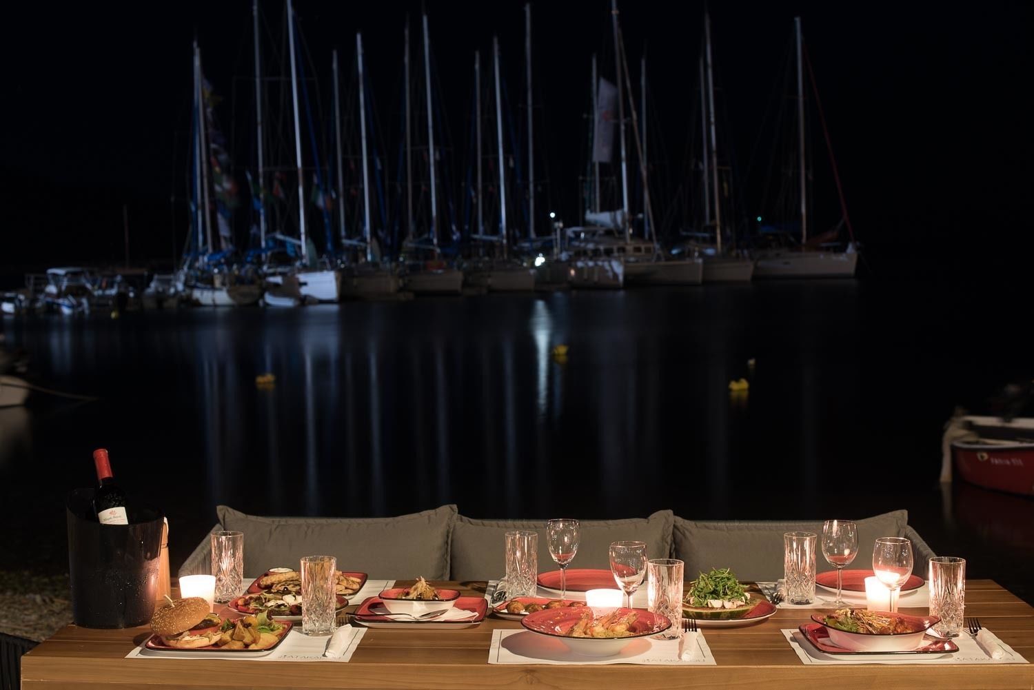 Une table dressée pour le dîner, avec assiettes et verres, donnant sur une marina où sont amarrés des bateaux.