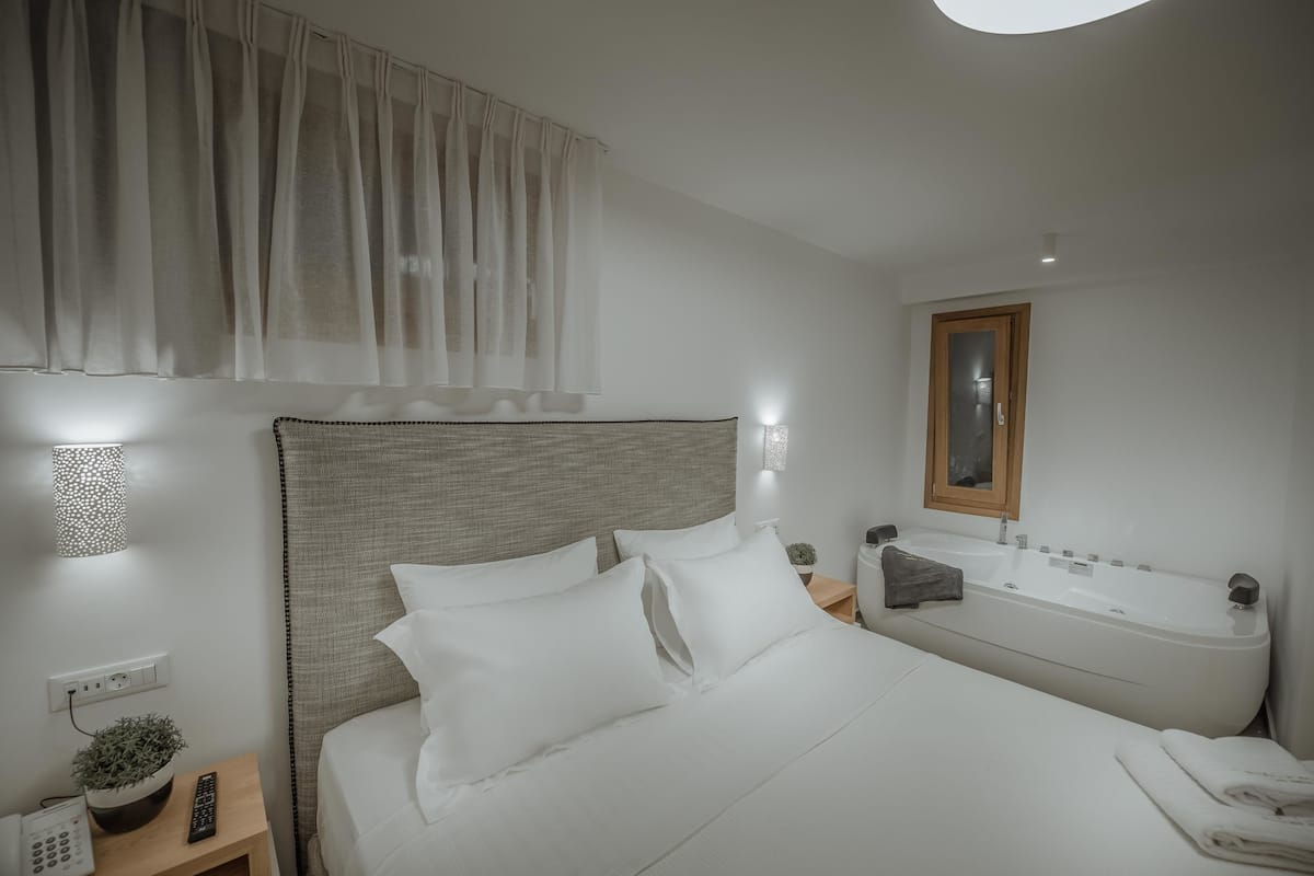 Une chambre moderne avec un grand lit, une table de chevet avec une lampe, une petite plante et une salle de bain avec une baignoire et un miroir.